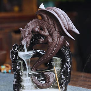 Könnte beinhalten: Ein brauner Drachen-Räuchergefäß mit Wasserfalleffekt. Der Drache sitzt auf einem dekorativen schwarzen Sockel, wobei Rauch herabfällt. Ein weißer Räucherkegel befindet sich auf dem Rücken des Drachen.