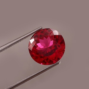 Puede incluir: Una gema de rubí redonda y facetada sostenida por unas pinzas. La gema es de color rojo intenso con una superficie brillante y reflectante. Las facetas crean un efecto de estallido estelar, resaltando el brillo de la gema. Las pinzas son plateadas.