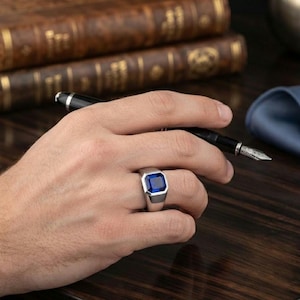 Puede incluir: Un anillo de tono plateado con una gran piedra preciosa de color azul intenso y corte cuadrado engastada en una banda de metal pulido. El anillo tiene un cuerpo ancho y sólido que se estrecha ligeramente hacia abajo, con un diseño geométrico.