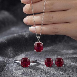 Könnte beinhalten: Ein Schmuckset bestehend aus einer silbernen Halskette mit einem kissenförmigen roten Edelsteinanhänger, einem passenden silbernen Ring mit einem kissenförmigen roten Edelstein und zwei silbernen Ohrsteckern mit kissenförmigen roten Edelsteinen.