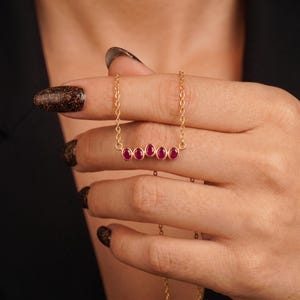 Può includere: Una collana a catena dorata con un pendente a barra orizzontale incastonato con cinque piccole pietre preziose ovali di un rosso rosato intenso. La sottile catena metallica si collega a entrambi i lati della montatura centrale, creando un gioiello raffinato e simmetrico.
