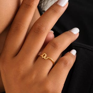 May include: A thin gold ring featuring a lowercase letter 'a' as the central design element. The band is smooth and circular, with the letter 'a' formed from the same continuous gold wire, creating a delicate and minimalist appearance on the finger.