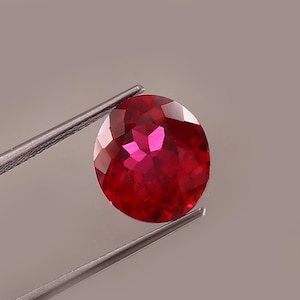 Op de afbeelding: Een gefacetteerde, ovaalvormige robijn edelsteen, vastgehouden door een pincet. De robijn heeft een dieprode kleur met meerdere facetten die licht reflecteren. De pincet is zilverkleurig en houdt de edelsteen tegen een neutrale achtergrond.