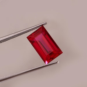 Könnte beinhalten: Ein rechteckiger, facettierter Rubin, der von einer silbernen Pinzette gehalten wird. Der Rubin hat eine tiefe, leuchtende rote Farbe mit sichtbaren Innenfacetten. Der Edelstein befindet sich vor einem neutralen Hintergrund.