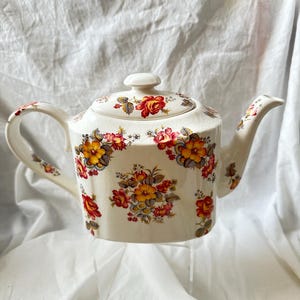 May include: Cream-colored ceramic teapot with a lid, handle, and spout. The teapot is decorated with a floral pattern featuring red, yellow, and orange roses. The design is repeated on the lid and handle. A classic piece for tea lovers.