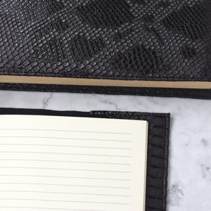 Puede incluir: Dos cuadernos negros con páginas rayadas. Las cubiertas tienen un patrón texturizado similar a la piel de serpiente. Un cuaderno está abierto, revelando las páginas interiores, mientras que el otro está cerrado. Las páginas son de color crema.