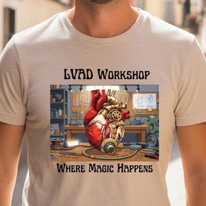 Könnte beinhalten: Beigefarbenes T-Shirt mit einem detaillierten Grafikdesign eines mechanischen Herzens in einer Werkstatt. Die Texte "LVAD WORKSHOP" und "WHERE MAGIC HAPPENS" sind oberhalb und unterhalb des Bildes aufgedruckt.