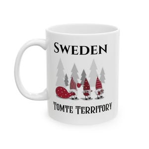 May include: White ceramic mug with the word "SWEDEN" in black, above a graphic of gnomes and trees. The text "TOMTE TERRITORY" is below the graphic. The gnomes are wearing red and white hats and carrying gifts.