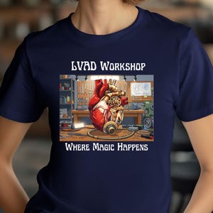 Könnte beinhalten: Marineblaues T-Shirt mit einem detaillierten Grafikdesign eines mechanischen Herzens in einer Werkstatt. Der Text "LVAD WORKSHOP" und "WHERE MAGIC HAPPENS" sind über und unter dem Bild zu sehen.