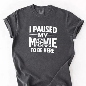 Puede incluir: Camiseta gris oscuro con mangas enrolladas. El texto blanco dice "I PAUSED MY MOVIE TO BE HERE". La palabra "MOVIE" incluye un gráfico de carrete de película. Una camiseta informal y cómoda.