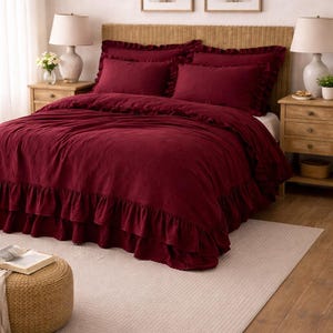 May include: A bedroom scene with a bed dressed in a deep red duvet cover and matching pillows, with a ruffled edge. The bed is on a light beige rug, with wooden bedside tables. Two framed floral prints hang above the bed.