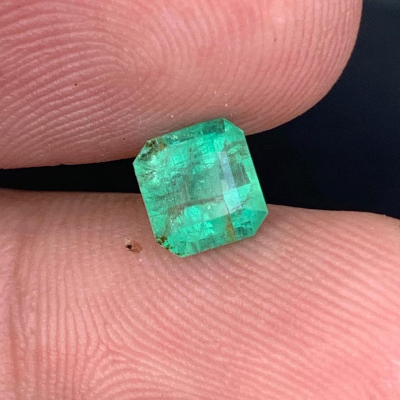 May include: A vibrant, emerald-cut emerald gemstone. The gem is a vivid green color with visible internal features and facets. The emerald is held between fingers, showcasing its clarity and brilliance.