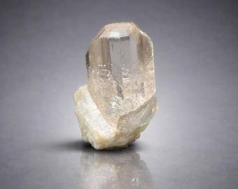 May include: A translucent, peach-colored crystal with a faceted, elongated shape. The crystal sits atop a rough, off-white mineral base. The lighting highlights the crystal's internal structure and clarity, creating a captivating display of natural beauty.