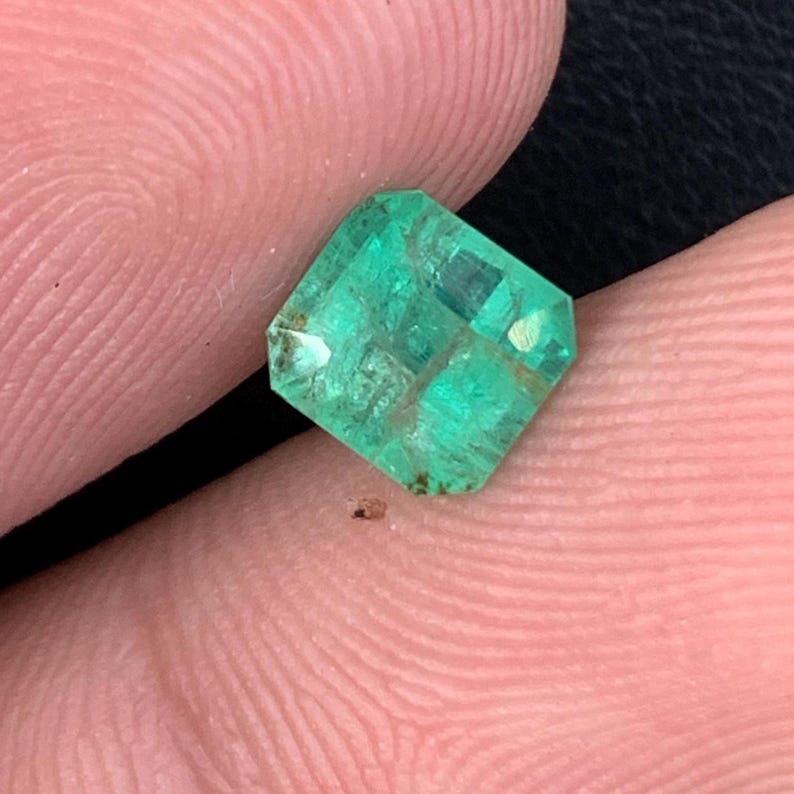 May include: A square-cut, vibrant green emerald gemstone. The gem has a faceted surface, reflecting light. The emerald is held between fingers, showcasing its clarity and color. The gemstone is a rich, deep green.