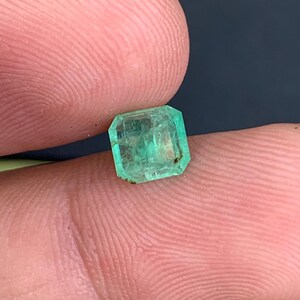 May include: A small, emerald-cut emerald gemstone. The gem is a vibrant green color with visible internal inclusions. The emerald is held between two fingers, showcasing its size and shape. The gemstone has a rectangular shape with beveled edges.