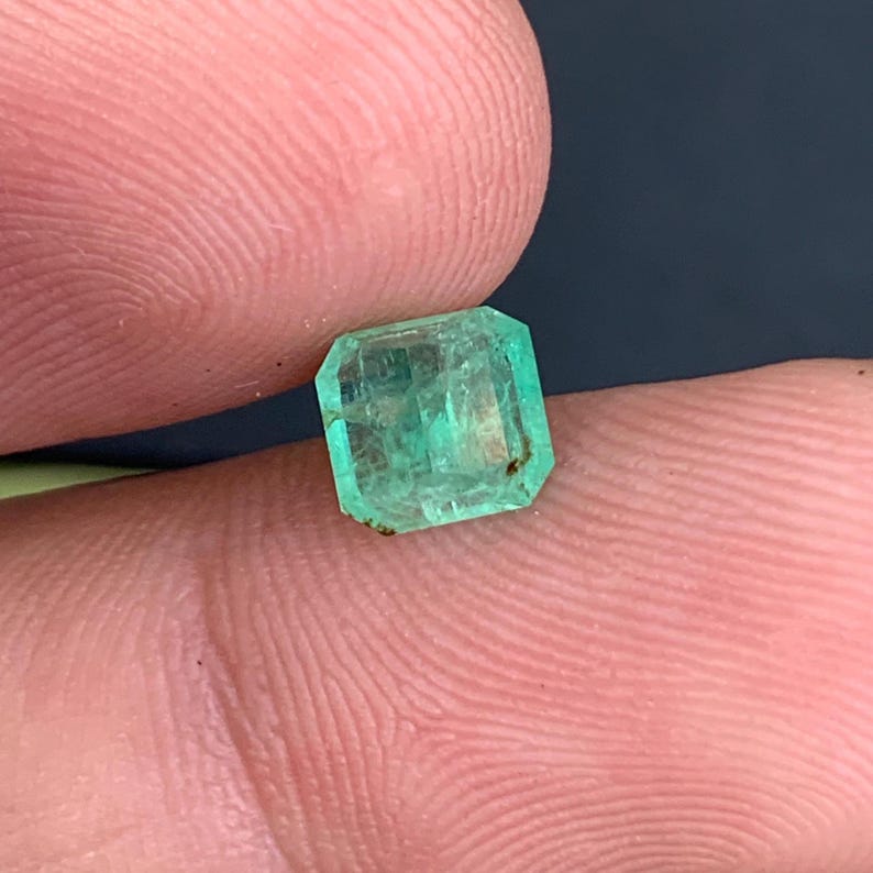 May include: A small, emerald-cut emerald gemstone. The gem is a vibrant green color with visible internal inclusions. The emerald is held between two fingers, showcasing its size and shape. The gemstone has a rectangular shape with beveled edges.
