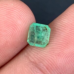 May include: A faceted, emerald-cut emerald gemstone. The gem is a vibrant green color with visible internal inclusions. The emerald is held between two fingers against a dark background, showcasing its clarity and color.