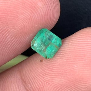 May include: A vibrant, emerald-cut green gemstone with visible internal features. The gem is held between two fingers, showcasing its clarity and color. The stone has a rectangular shape with beveled edges, reflecting light.