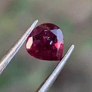 Puede incluir: Un rubí facetado en forma de corazón, sostenido por unas pinzas plateadas. El rubí muestra un color rojo intenso y superficies reflectantes. El fondo es verde borroso, resaltando la belleza de la gema.