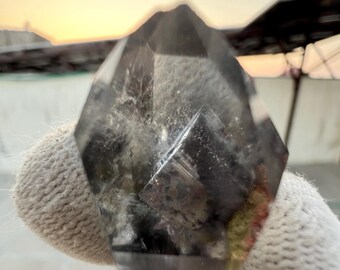 May include: A faceted, smoky quartz crystal with a dark, translucent appearance. The crystal has a pointed top and multiple geometric faces. The color transitions from a dark gray to a lighter, almost clear hue. The crystal is held by a gloved hand.