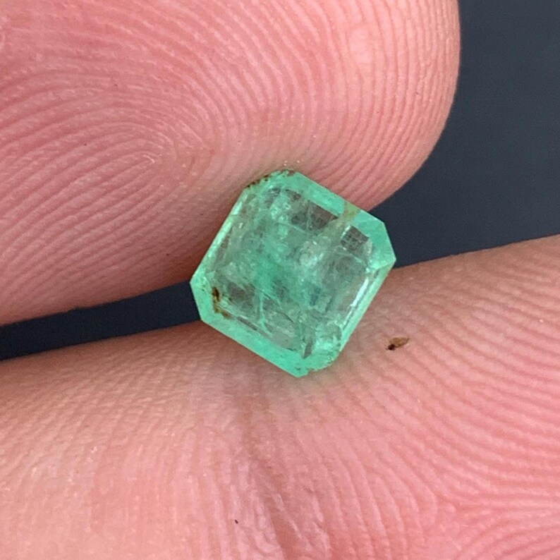 May include: A faceted, emerald-cut emerald gemstone. The gem is a vibrant green color with visible internal inclusions. The emerald is held between two fingers, showcasing its size and clarity. The cut features multiple facets.