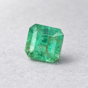 May include: A faceted emerald gemstone with a rectangular shape. The gem is a vibrant green color with visible inclusions and a polished surface. The emerald is set against a neutral background.