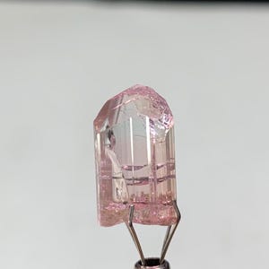 Peut inclure: Un cristal de tourmaline rose clair translucide avec une structure prismatique verticale allongée. La pierre précieuse présente des stries naturelles sur toute sa longueur, une terminaison facettée au sommet et des inclusions internes visibles.