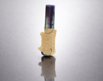 May include: A close-up of a tourmaline crystal. The crystal is a deep purple at the top, transitioning to green and then a clear base. It is partially encased in a rough, beige matrix. The crystal is standing upright.