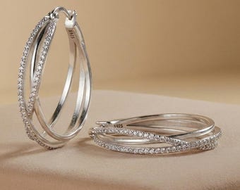 May include: A pair of sterling silver hoop earrings featuring a triple-band crossover design. Each band is encrusted with small clear stones, and the metal is stamped with S925. One earring stands upright while the other lies flat.