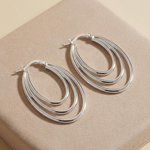 May include: A pair of silver-toned hoop earrings, each featuring three interconnected oval-shaped loops. The earrings have a polished, reflective metallic finish and include a hinged post mechanism for secure closure at the top of the hoops.