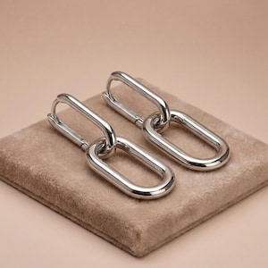 May include: A pair of silver-coloured chain link earrings. Each earring has two elongated oval links connected. The earrings are displayed on a square, tan, plush surface against a neutral background.
