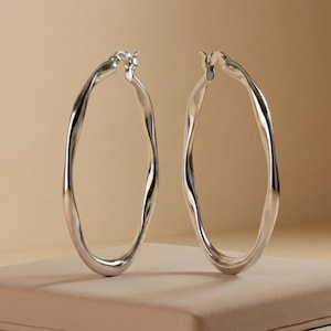 May include: A pair of polished silver hoop earrings featuring a distinctively irregular, wavy, and organic silhouette. The metal surface is smooth and reflective, showcasing a continuous, non-uniform circular shape with a hinged closure mechanism.
