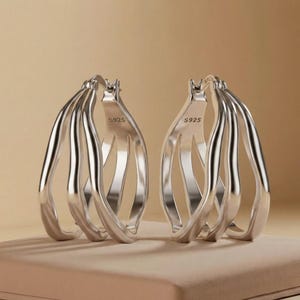 May include: A pair of silver hoop earrings featuring a multi-layered, wavy design with three distinct bands. Each earring is crafted from polished metal and includes the hallmark S925 stamped clearly on the inner surface of the hoop structure.