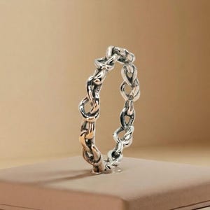 May include: A silver ring with a chain link design. The ring is made of a shiny metal and has a detailed, textured appearance. The chain links are intertwined, creating a continuous loop. The ring is displayed on a neutral-colored surface.