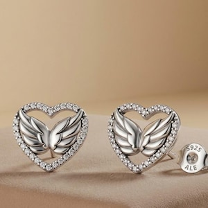May include: A pair of silver stud earrings. Each earring features a heart-shaped design with a border of small, clear crystals. Inside the heart is a detailed wing design. One earring post is visible, stamped with "925 ALE".