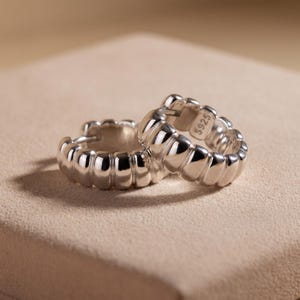 May include: A pair of silver hoop earrings with a ribbed, textured surface. The earrings feature a rounded, chunky design with a polished finish. One of the earrings is clearly stamped with the text S925 on the inner side of the metal band.
