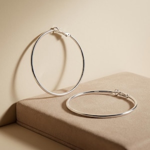 May include: A pair of silver hoop earrings with a smooth, polished finish. One earring stands upright while the other lies flat. Each hoop features a secure hinged clasp mechanism and is stamped with the 925 hallmark, indicating sterling silver.
