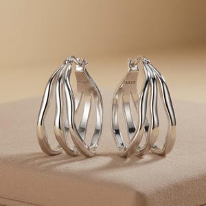 May include: A pair of silver hoop earrings featuring a triple-band design with a polished finish. Each earring is constructed from three interconnected, wavy metallic strands that form a continuous loop. The inner band is stamped with S925.