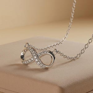 Può includere: Una collana color argento con un ciondolo a forma di simbolo dell'infinito. Il ciondolo è decorato sulla superficie con piccole pietre trasparenti a taglio rotondo. La catena è composta da delicati anelli circolari intrecciati.