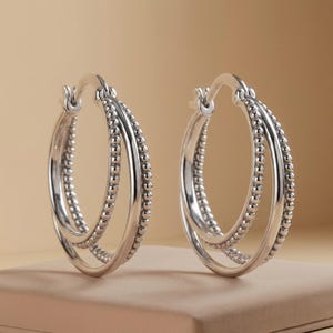 May include: A pair of silver hoop earrings featuring a double-loop design. Each earring consists of one smooth, polished silver band intertwined with a second band detailed with a continuous row of small, raised silver beads along the outer edge.