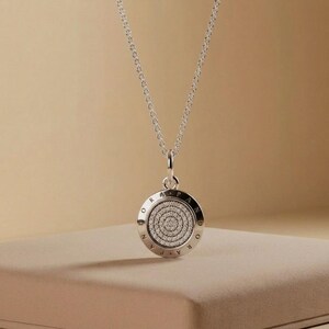 May include: A silver pendant necklace with a circular charm. The charm features a spiral of small, clear stones in the center, surrounded by a ring of text. The necklace chain is delicate and silver.