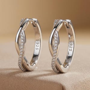 May include: A pair of silver hoop earrings featuring a twisted design with one side encrusted with small clear stones. The metal is stamped with S925, indicating sterling silver composition. The earrings have a polished finish and a hinged closure.