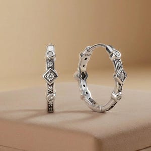 May include: A pair of silver hoop earrings featuring a repeating pattern of round clear stones and a central square-shaped stone setting. The metal is stamped with S925, indicating sterling silver composition, with a polished finish.