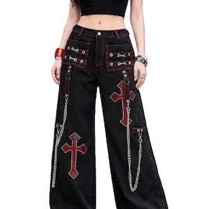 Op de afbeelding: Zwarte jeans met wijde pijpen, rode kruisborduurwerk en stiksels. De jeans hebben zilveren kettingaccenten, grommets en een ontwerp met hoge taille. Het model draagt een zwarte crop top.