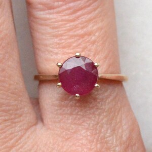 May include: A gold ring featuring a round, faceted, deep red gemstone set in a six-prong setting. The band is thin and polished, showcasing a classic design that highlights the vibrant color and circular cut of the central stone.