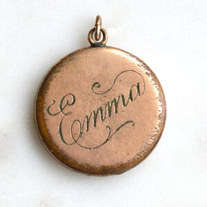 May include: A round, copper-toned pendant with the name "Emma" in elegant script. The pendant has a small loop at the top for a chain and shows signs of age, suggesting it is vintage or antique.