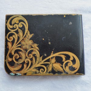 May include: A rectangular, black vintage compact with a gold floral design in the corner. The gold design includes flowers and swirling leaves. The compact has a slightly worn appearance.