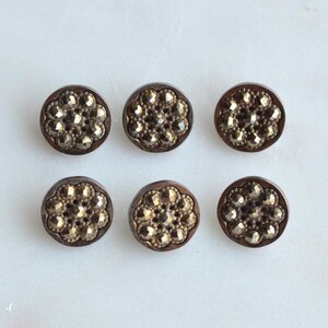 May include: Six antique, round buttons with a floral design. The buttons have a dark brown rim and a gold-colored, textured center. They are arranged in two rows of three on a white surface.
