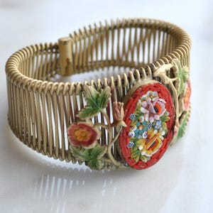 May include: A vintage gold-toned wire bracelet with a floral mosaic design. The oval mosaic features a bouquet of colorful flowers set against a red background. The bracelet is approximately 1.5 inches wide.