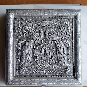May include: A silver-colored, rectangular box with an ornate embossed design. The central motif features two peacocks facing each other, surrounded by floral and foliate patterns. The box has a decorative border.